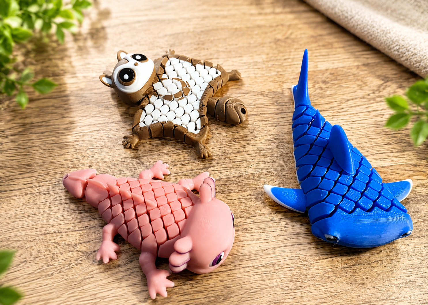 Three textured, flexi animal toys shaped like a squirrel, a pink axolotl, and a blue shark, arranged on a wooden surface with green leaves and a beige cloth in the background.